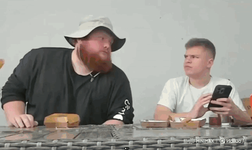 A Man With A Beard Wearing A Hat Is Sitting At A Table With Another Man . GIF