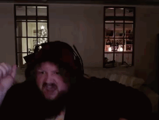 A Man With A Beard Wearing A Hooded Jacket Is Standing In A Dark Room With A Christmas Tree In The Background GIF