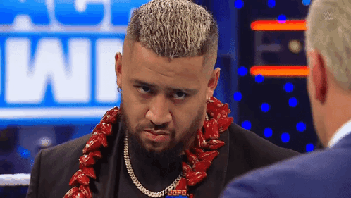A Man With A Beard Wearing A Lei Is Talking To Another Man In A Suit . GIF