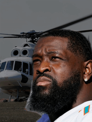 a man with a beard wearing a miami dolphins shirt stands in front of a helicopter