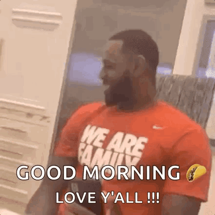 A Man With A Beard Wearing A Red Shirt That Says `` We Are Family '' Is Sitting In Front Of A Window . GIF
