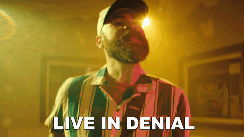 A Man With A Beard Wearing A Striped Shirt And A Hat Says Live In Denial GIF