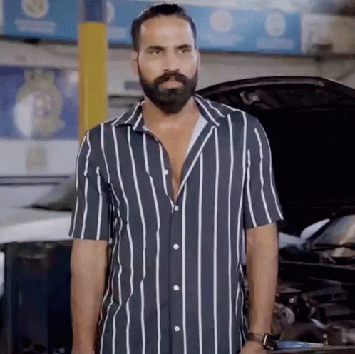 A Man With A Beard Wearing A Striped Shirt Stands In Front Of A Car GIF