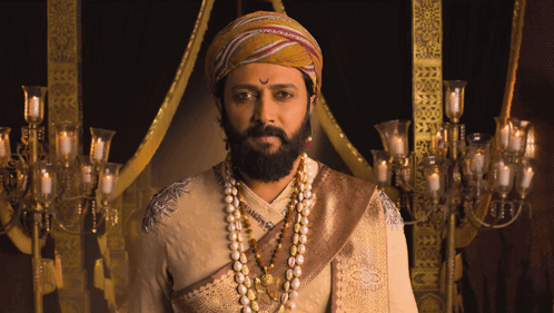 A Man With A Beard Wearing A Turban And Pearls Looks At The Camera GIF