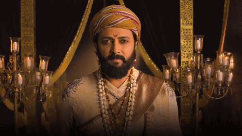 A Man With A Beard Wearing A Turban And Pearls Looks At The Camera GIF