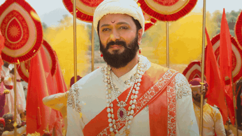 A Man With A Beard Wearing A Turban And Pearls Stands In Front Of Red Flags GIF