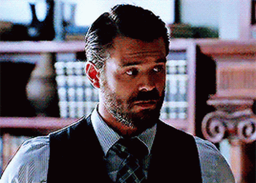 A Man With A Beard Wearing A Vest And Tie Stands In Front Of A Bookshelf GIF