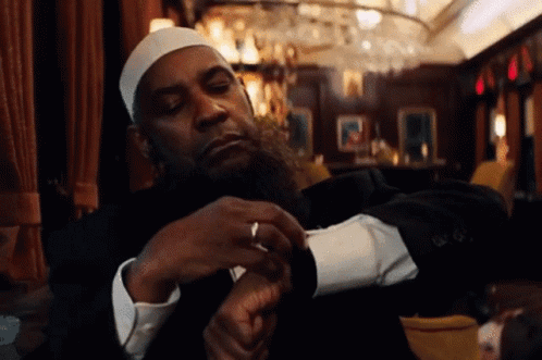 A Man With A Beard Wearing A White Hat And A Black Suit Is Looking At His Watch GIF