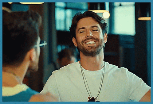A Man With A Beard Wearing A White T-Shirt Smiles While Talking To Another Man GIF