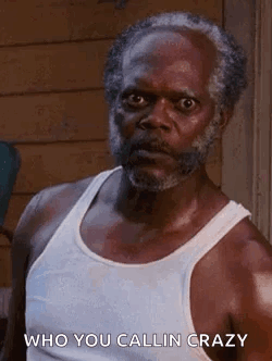 A Man With A Beard Wearing A White Tank Top Is Standing In Front Of A Building . GIF