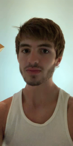A Man With A Beard Wearing A White Tank Top Looks At The Camera GIF