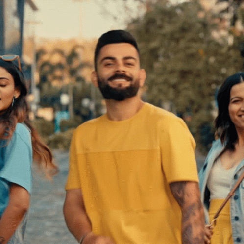 A Man With A Beard Wearing A Yellow T-Shirt Smiles With Two Women Behind Him GIF