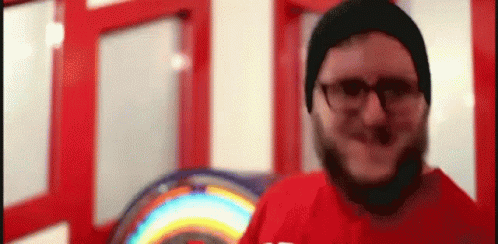 A Man With A Beard Wearing Glasses And A Beanie Is Standing In Front Of A Rainbow Sign . GIF