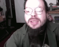 A Man With A Beard Wearing Glasses And A Green Jacket Is Talking . GIF
