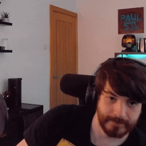 A Man With A Beard Wearing Headphones In Front Of A Paul Stan Poster GIF