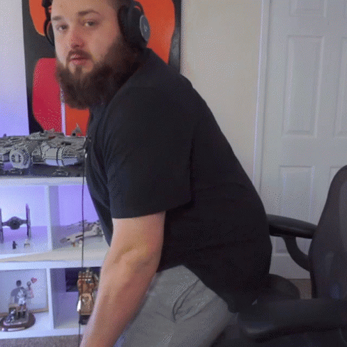 A Man With A Beard Wearing Headphones Is Sitting In Front Of A Shelf With Toys On It GIF