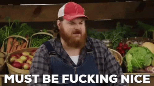A Man With A Beard Wearing Overalls And A Hat Is Standing In Front Of Baskets Of Fruit And Vegetables . GIF