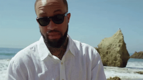 A Man With A Beard Wearing Sunglasses And A White Shirt Stands On The Beach GIF
