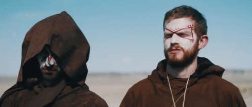 A Man With A Cross Painted On His Face Stands Next To Another Man With A Hood GIF