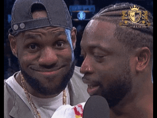 A Man With A Crown On His Head Talks Into A Microphone Next To Another Man GIF