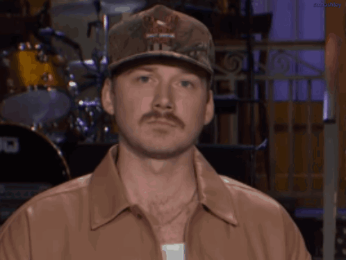 A Man With A Hat And A Mustache Is Wearing A Brown Shirt GIF