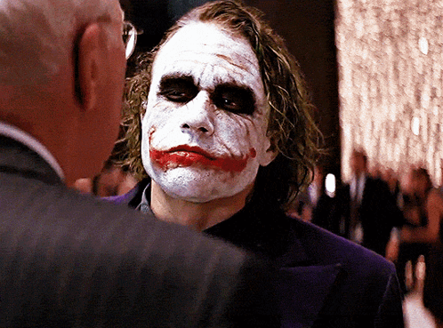 A Man With A Joker Face Painted On His Face Talks To Another Man In A Suit GIF
