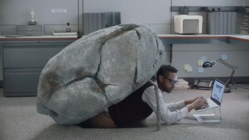 A Man With A Large Rock On His Back Is Using A Laptop In An Office GIF