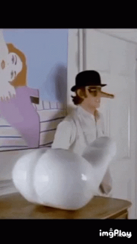 A Man With A Long Nose And A Hat Is Standing Next To A Large White Object On A Table . GIF