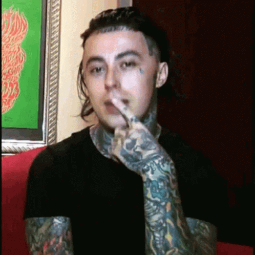A Man With A Lot Of Tattoos On His Arms Is Sitting On A Couch With His Finger In His Mouth . GIF