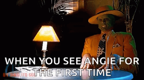 A Man With A Mask On His Face Is Sitting At A Table With A Lamp And Says When You See Angie For The First Time GIF