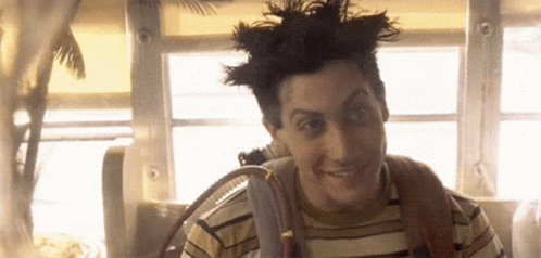 A Man With A Mohawk And A Striped Shirt Is Smiling While Sitting In Front Of A Window . GIF