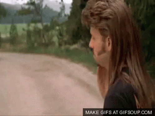 A Man With A Mohawk And Long Hair Is Standing On A Road . GIF
