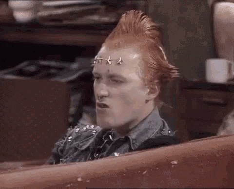 A Man With A Mohawk And Studs On His Head Is Sitting On A Couch And Making A Funny Face . GIF
