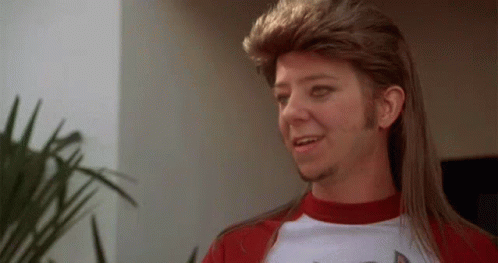 a man with a mullet and a beard is wearing a red shirt .