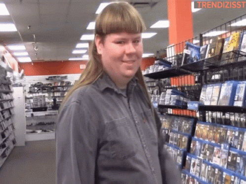 A Man With A Mullet Is Standing In Front Of A Shelf Of Dvds GIF