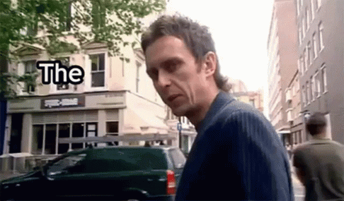 A Man With A Mullet Is Walking Down A Street In Front Of A Building With The Word The On It . GIF