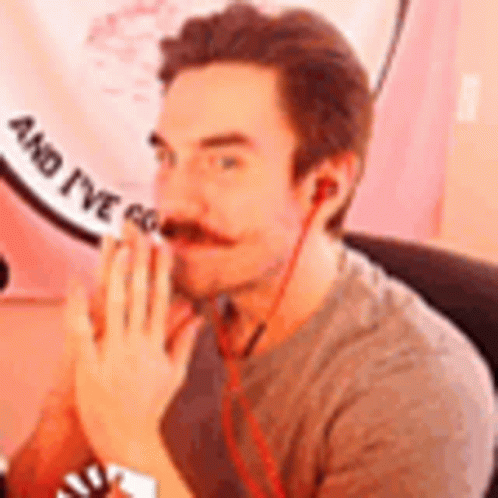 A Man With A Mustache And Ear Buds Is Sitting In Front Of A Pink Sign . GIF