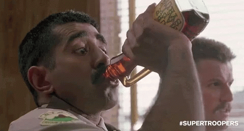 a man with a mustache is drinking from a bottle with a label that says ' supertroopers ' on it .