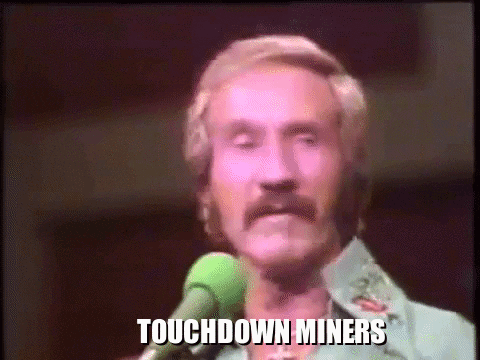 a man with a mustache is singing into a microphone with the words touchdown miners written on the screen behind him .