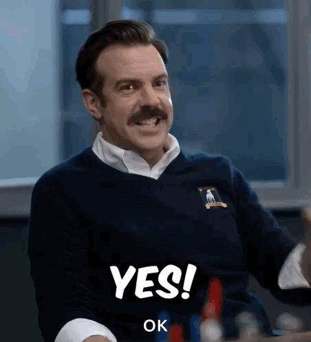 A Man With A Mustache Is Sitting At A Table And Smiling And Saying Yes . GIF