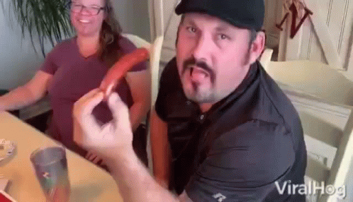 A Man With A Mustache Is Sitting At A Table With A Woman Holding A Hot Dog . GIF