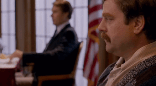 A Man With A Mustache Is Sitting In A Chair In Front Of A Flag . GIF