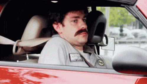 A Man With A Mustache Is Sitting In A Red Car Looking Out The Window . GIF