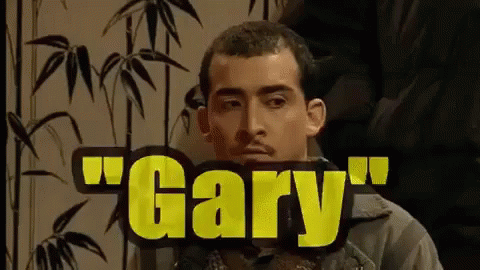 A Man With A Mustache Is Sitting In Front Of A Sign That Says Gary . GIF
