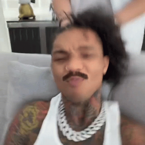 A Man With A Mustache Is Sitting On A Couch With His Hair In A Ponytail . GIF