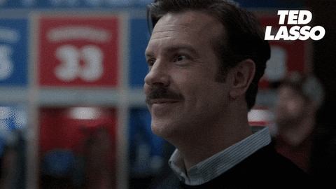 A Man With A Mustache Is Smiling In Front Of A Ted Lasso Sign GIF