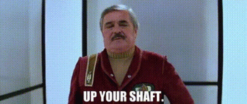 a man with a mustache is standing in a hallway with the words `` up your shaft '' written on the screen .