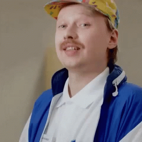 A Man With A Mustache Is Wearing A Blue Jacket And A Hat . GIF