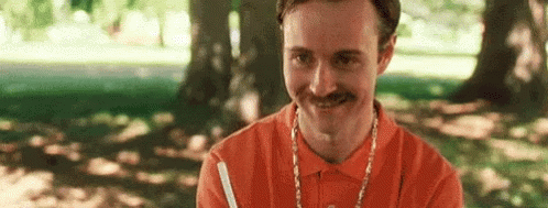 A Man With A Mustache Is Wearing An Orange Polo Shirt And A Necklace . GIF