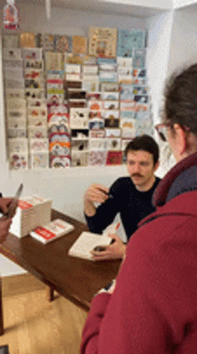 A Man With A Mustache Sits At A Table In Front Of A Wall Full Of Cards GIF